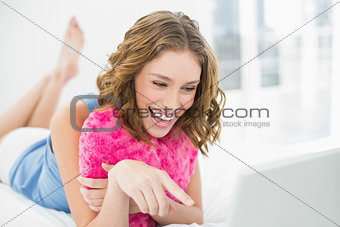 Lovely laughing woman cuddling with a heart pillow pointing at her notebook