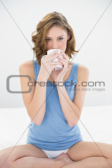 Calm attractive woman sitting on her bed drinking from white cup