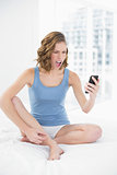 Angry beautiful woman sitting on her bed looking at her smartphone