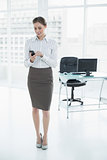 Gorgeous calm businesswoman standing in her office using her smartphone