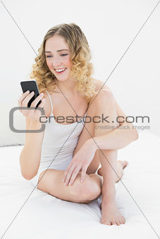 Pretty happy blonde sitting on bed holding smartphone