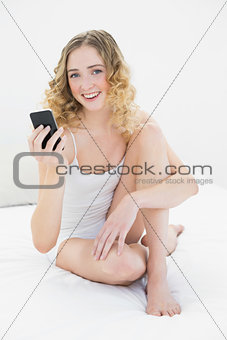 Pretty smiling blonde sitting on bed holding smartphone