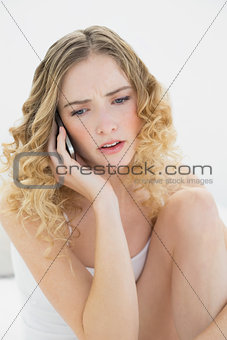 Pretty upset blonde sitting on bed phoning
