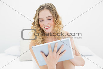 Pretty smiling blonde sitting on bed using tablet