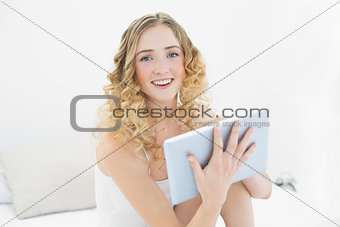 Pretty cheerful blonde sitting on bed using tablet