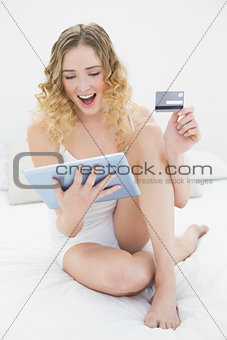 Pretty excited blonde sitting on bed holding tablet and credit card