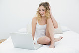 Pretty smiling blonde sitting on bed using laptop