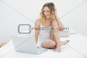 Pretty smiling blonde sitting on bed using laptop