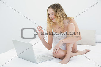 Pretty smiling blonde sitting on bed waving at laptop