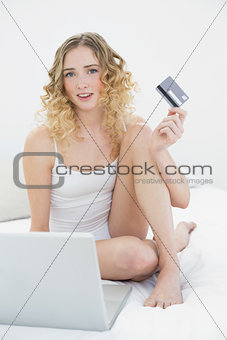 Pretty happy blonde sitting on bed using laptop and credit card