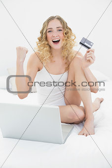 Pretty successful blonde sitting on bed using laptop and credit card