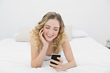 Pretty smiling blonde lying on bed using smartphone