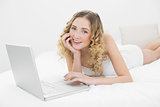 Pretty smiling blonde lying on bed using laptop