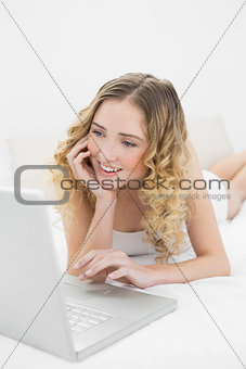 Pretty cheerful blonde lying on bed using laptop
