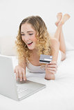Pretty excited blonde lying on bed using laptop for online shopping