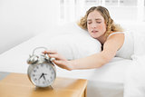 Pretty tired blonde lying in bed reaching for alarm clock