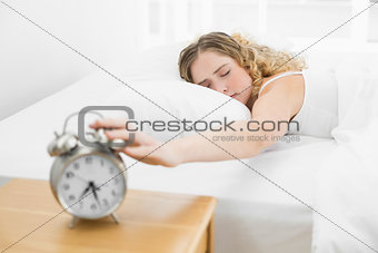 Pretty tired blonde lying in bed turning off alarm clock