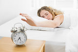 Pretty upset blonde lying in bed reaching for alarm clock