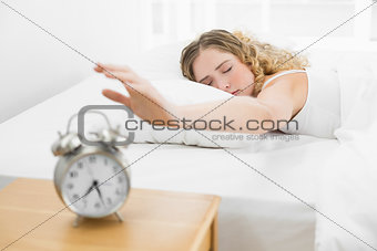 Pretty upset blonde lying in bed reaching for alarm clock