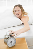 Pretty upset blonde lying in bed turning off alarm clock