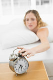 Pretty annoyed blonde lying in bed turning off alarm clock