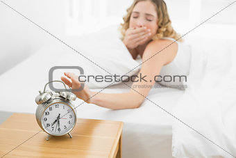 Pretty yawning blonde lying in bed turning off alarm clock