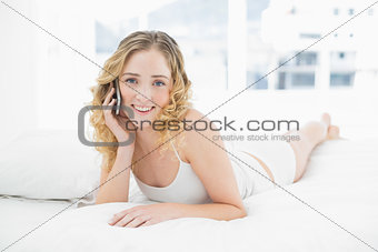 Pretty smiling blonde lying in bed phoning