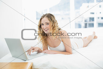 Pretty cheerful blonde lying in bed using laptop