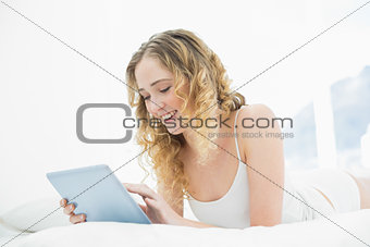 Pretty smiling blonde lying in bed using tablet