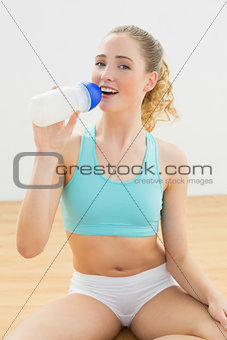 Smiling slim blonde sitting on floor drinking from sports bottle