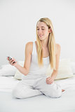 Attractive blonde woman sitting on her bed holding her smartphone