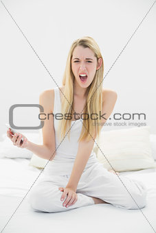 Shocked blonde woman holding her smartphone while sitting on her bed