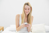 Content blonde woman texting with her smartphone sitting on her bed