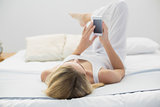 Blonde young woman using her smartphone while lying on her bed