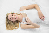Happy blonde woman using her smartphone lying on her bed