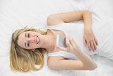 Joyful blonde woman holding her smartphone lying on her bed