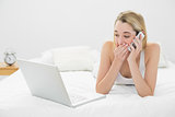 Beautiful shocked woman phoning while lying on her bed looking at laptop