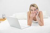 Young surprised woman phoning while lying on her bed next to her notebook