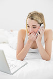 Blonde shocked woman phoning and using her notebook lying on her bed
