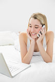 Smiling blonde woman lying on her bed phoning using her notebook