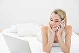 Beautiful woman phoning while lying on her bed using her notebook