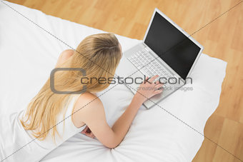 Slender blonde woman using her notebook lying on her bed