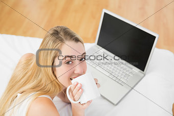 Lovely woman lying on bed next to laptop drinking of cup
