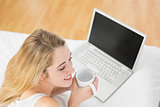 Calm blonde woman holding a cup lying on bed next to her laptop
