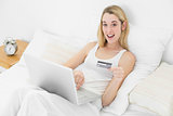 Astonished young woman using her laptop for home shopping while looking at camera