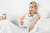 Happy blonde woman using her laptop for home shopping lying on her bed