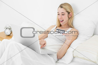 Happy blonde woman using her laptop for home shopping lying on her bed