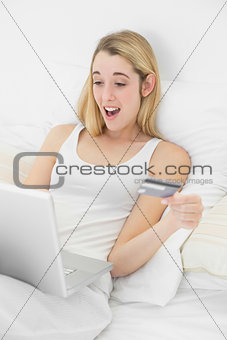 Happy surprised woman home shopping with her laptop lying on her bed