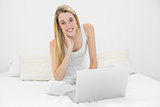Content blonde woman using her notebook sitting on her bed smiling at camera