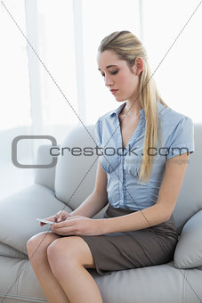 Serious beautiful businesswoman texting with her smartphone sitting on couch